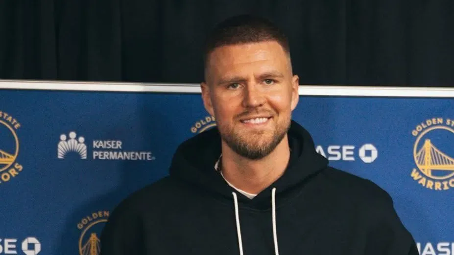 Homem sorridente de moletom preto em frente a painel azul com logos do Golden State Warriors e da Kaiser Permanente