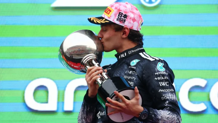 Piloto de macacão preto da Mercedes beija um troféu prateado no pódio, usando boné rosa com estampa floral e a inscrição "日本 2026