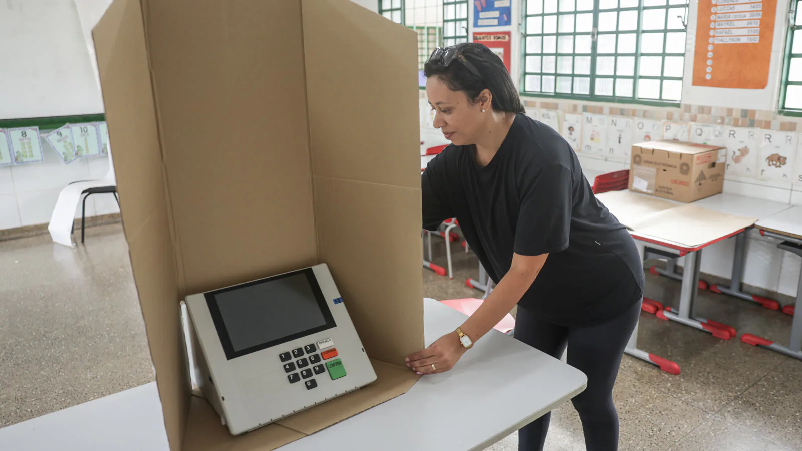 Mesária ajeita a cabine de papelão ao redor de uma urna eletrônica em uma sala de aula de escola pública em Brasília, em 24 de outubro de 2026 (imagem de arquivo, não é do exterior)