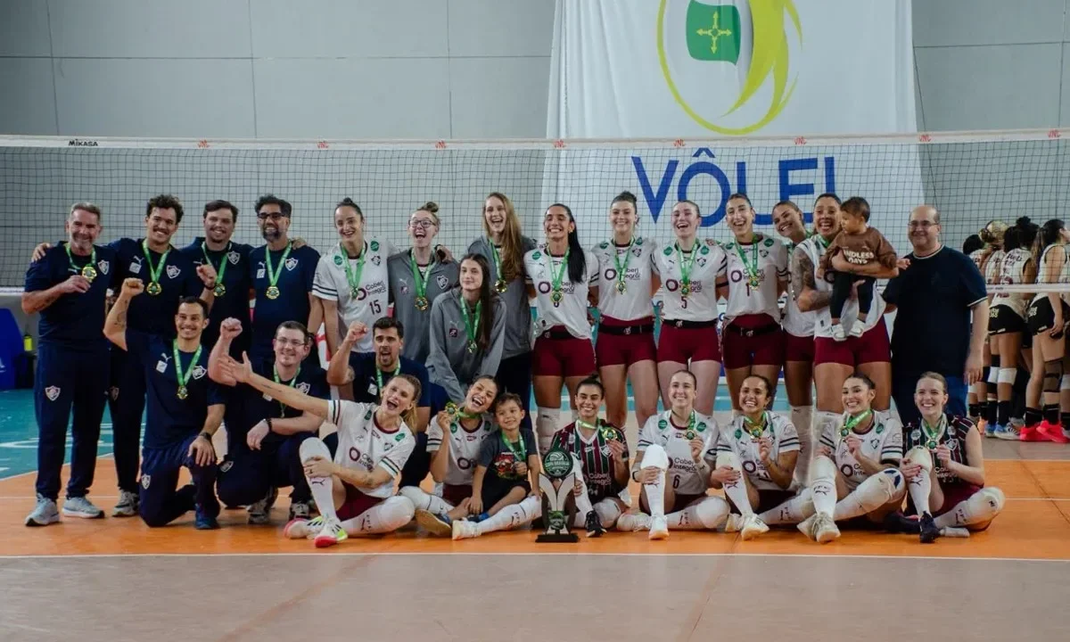 Equipe de vôlei feminino posa para foto com medalhas e troféu após conquista do título em competição realizada em ginásio coberto