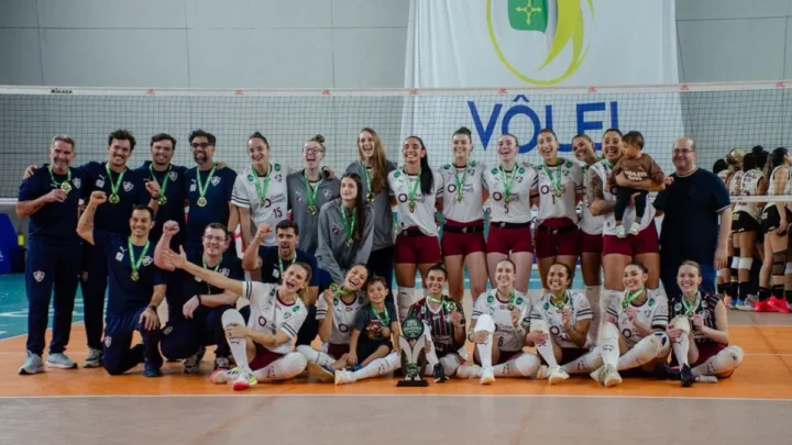 Equipe de vôlei feminino posa para foto com medalhas e troféu após conquista do título em competição realizada em ginásio coberto