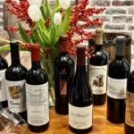 Garrafas de vinho tinto dispostas em fileira sobre superfície de madeira, com arranjo floral de tulipas brancas e frutas vermelhas ao fundo e parede de tijolos