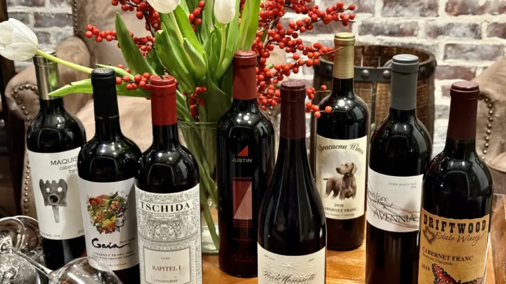 Garrafas de vinho tinto dispostas em fileira sobre superfície de madeira, com arranjo floral de tulipas brancas e frutas vermelhas ao fundo e parede de tijolos