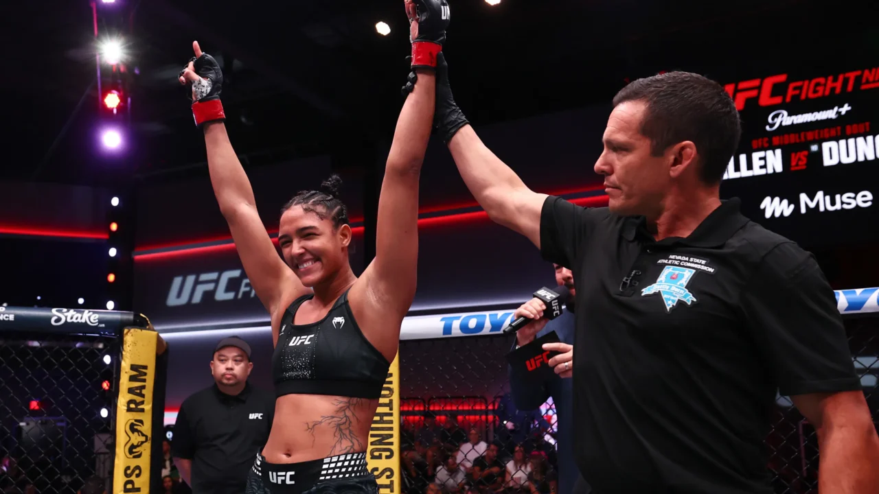 Lutadora de MMA com as mãos erguidas e sorriso largo ao lado de um árbitro de camisa preta da comissão atlética de Nevada, dentro do octógono