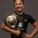Lutadora sorri enquanto segura cinturão hexagonal de ouro e preto, usando uniforme de combate preto com patrocínios