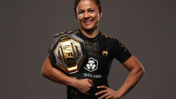 Lutadora sorri enquanto segura cinturão hexagonal de ouro e preto, usando uniforme de combate preto com patrocínios