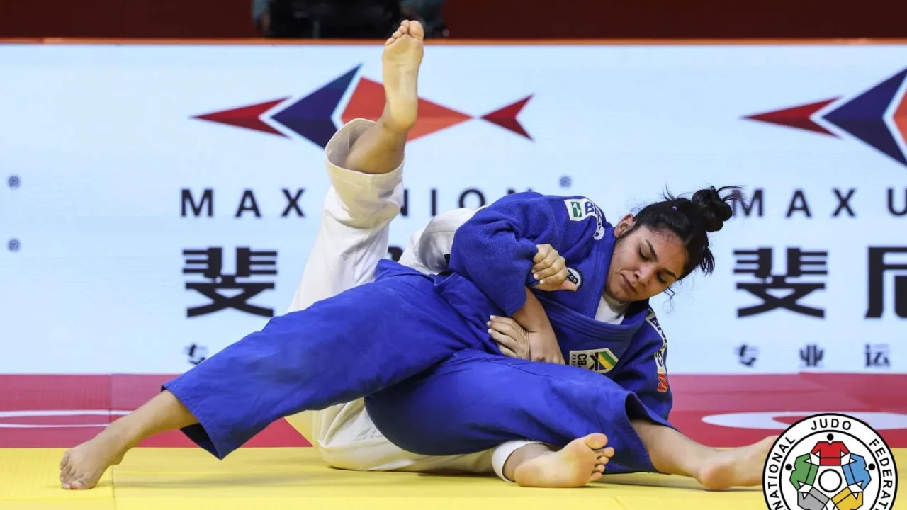 Judoca de faixa azul, com uniforme do Brasil, aplica uma técnica de chão sobre a adversária de branco em tatame amarelo, sob painel do Max Union