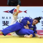 Judoca de uniforme azul com faixa do Brasil aplica golpe no chão sobre adversária de branco durante disputa em tatame amarelo