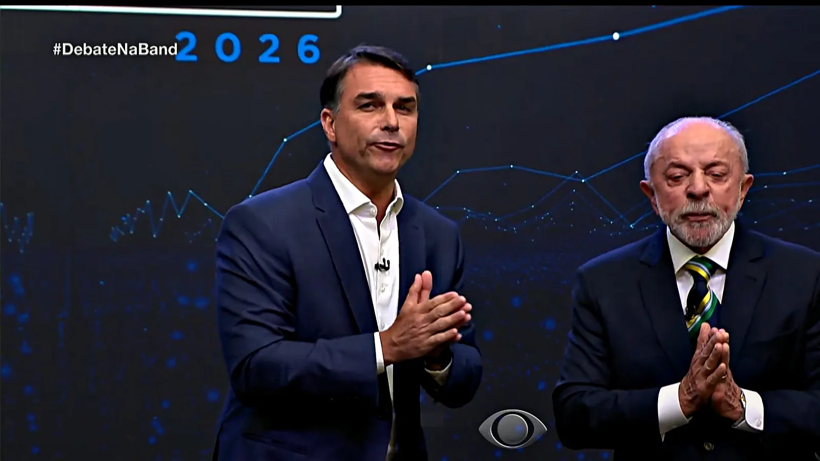 Flávio Bolsonaro fala de mãos juntas, ao lado de Lula, no palco do debate da Band