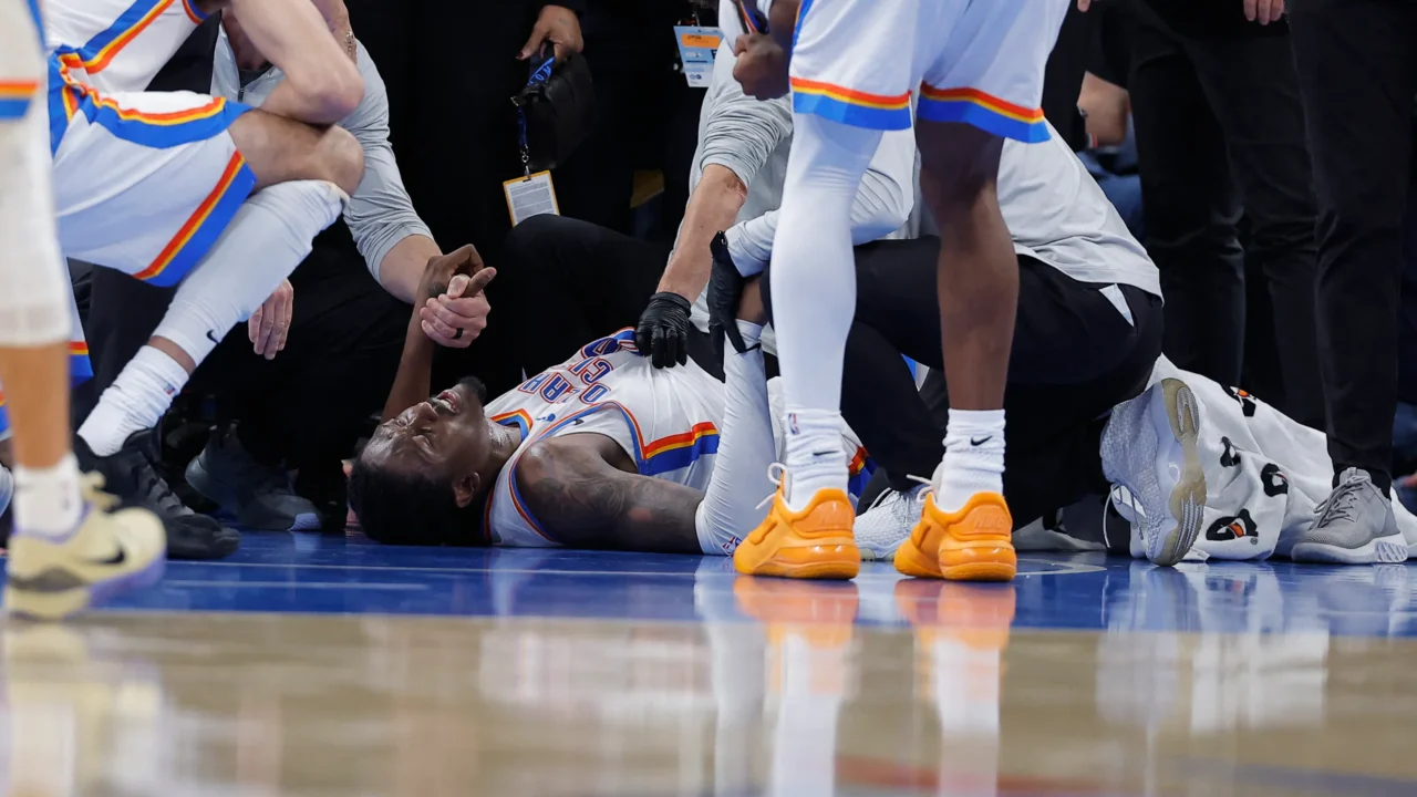 Jogador de camisa branca do Oklahoma City Thunder deitado no chão da quadra, com dor no rosto, cercado por companheiros e membros da equipe médica