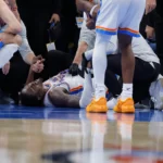 Jogador de camisa branca do Oklahoma City Thunder deitado no chão da quadra, com dor no rosto, cercado por companheiros e membros da equipe médica