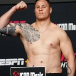 Homem sem camisa, com tatuagens no braço e no peito, flexiona o bíceps diante de painel com logos da ESPN e do UFC durante a pesagem