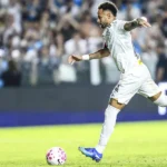 Jogador de camisa branca do Santos chuta a bola em campo, com torcida ao fundo e placa de publicidade ao lado do gramado
