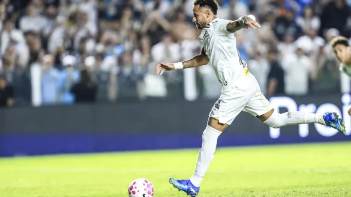 Jogador de camisa branca do Santos chuta a bola em campo, com torcida ao fundo e placa de publicidade ao lado do gramado