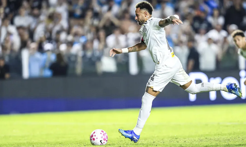 Jogador de camisa branca do Santos chuta a bola em campo, com torcida ao fundo e placa de publicidade ao lado do gramado