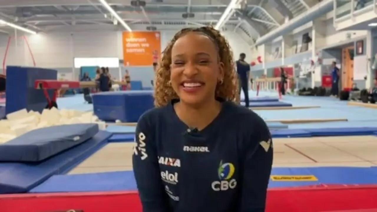 Atleta sorridente, com uniforme da CBG, em um ginásio de ginástica com colchões azuis e aparelhos ao fundo