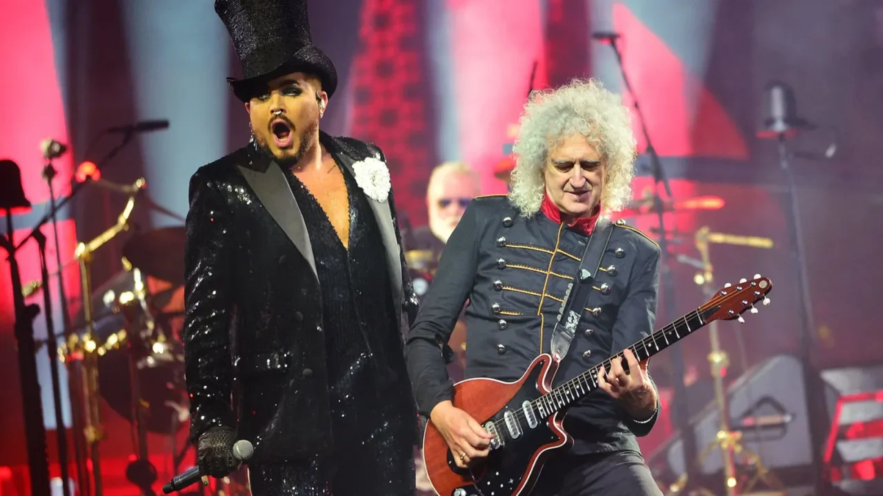 Apresentador em jaqueta com paetês e chapéu alto canta enquanto guitarrista com cabelo branco e casaco militar toca ao fundo em palco iluminado em tons de rosa e vermelho