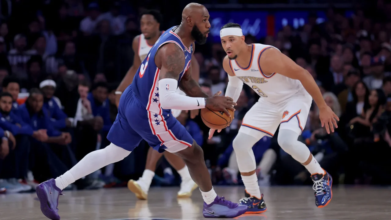 Jogador de camisa azul com faixas vermelhas e brancas conduz a bola em quadra enquanto um adversário de uniforme branco do New York marca de perto