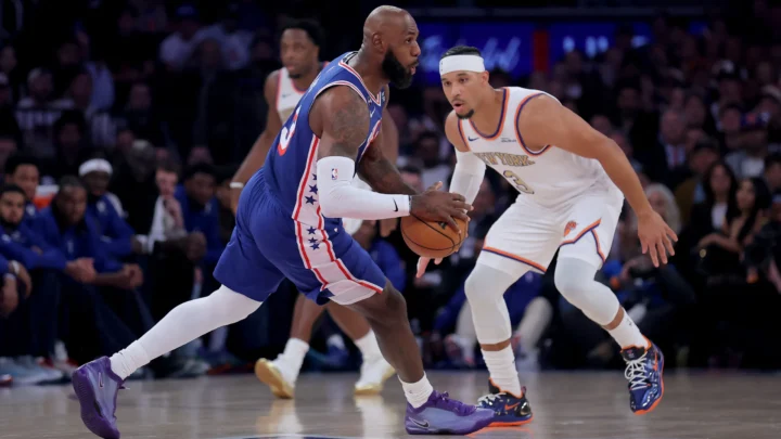 Jogador de camisa azul com faixas vermelhas e brancas conduz a bola em quadra enquanto um adversário de uniforme branco do New York marca de perto
