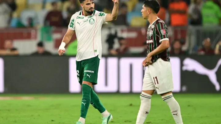 Jogador do Palmeiras, de camisa branca e calção verde, levanta o braço ao lado de um adversário de camisa listrada do Fluminense durante partida em campo