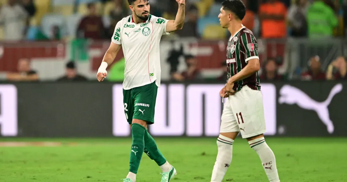Jogador do Palmeiras, de camisa branca e calção verde, levanta o braço ao lado de um adversário de camisa listrada do Fluminense durante partida em campo