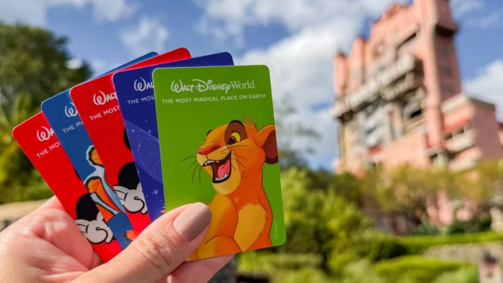 Mão segura ingressos de parque temático com a marca Disney e personagens em frente a edifício de alvenaria e céu azul