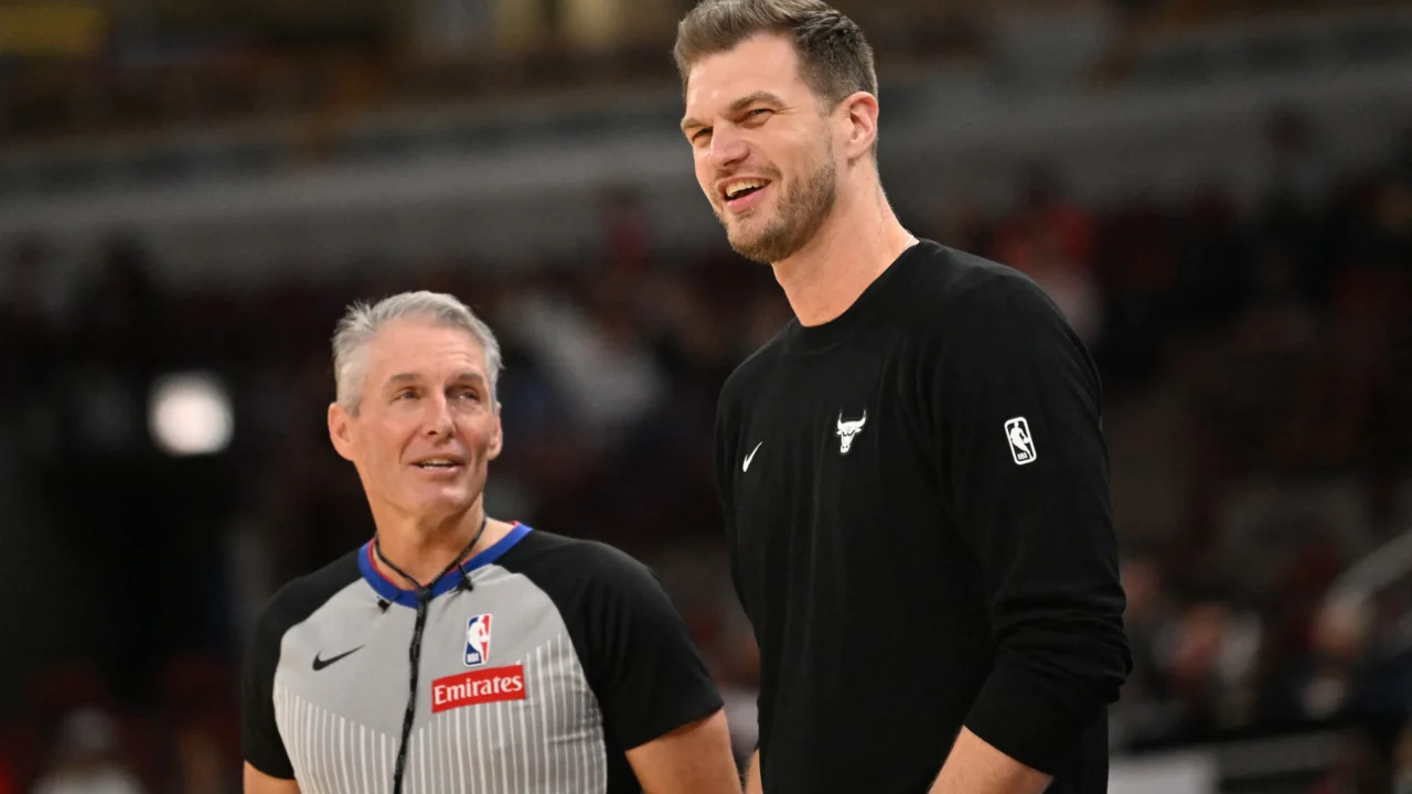 Um árbitro de cabelos grisalhos, de uniforme cinza e preto da NBA, conversa sorridente com um homem de suéter preto com o logo dos Bulls, em uma quadra de basquete