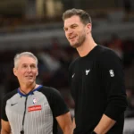 Um árbitro de cabelos grisalhos, de uniforme cinza e preto da NBA, conversa sorridente com um homem de suéter preto com o logo dos Bulls, em uma quadra de basquete