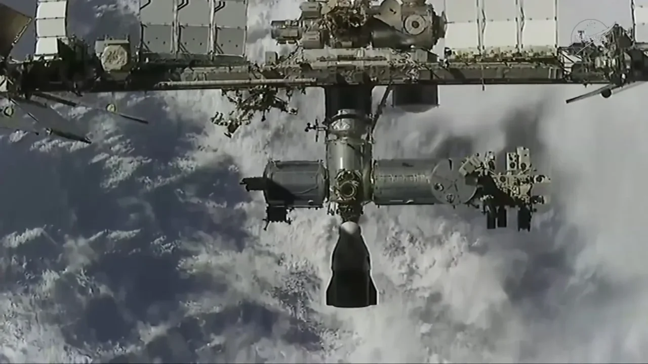 A nave Dragon, pequena e escura contra as nuvens da Terra, se aproxima por baixo da Estação Espacial Internacional