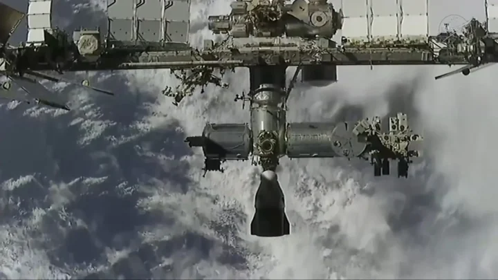 A nave Dragon, pequena e escura contra as nuvens da Terra, se aproxima por baixo da Estação Espacial Internacional