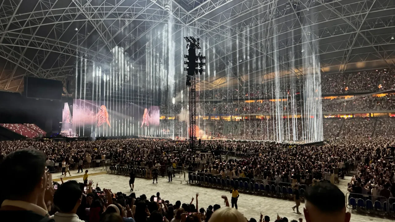 Apresentação de espetáculo em estádio coberto com estrutura metálica, palco central iluminado por feixes de luz verticais brancos, multidão nas arquibancadas e público próximo ao piso da arena acompanhando o show