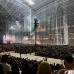 Apresentação de espetáculo em estádio coberto com estrutura metálica, palco central iluminado por feixes de luz verticais brancos, multidão nas arquibancadas e público próximo ao piso da arena acompanhando o show