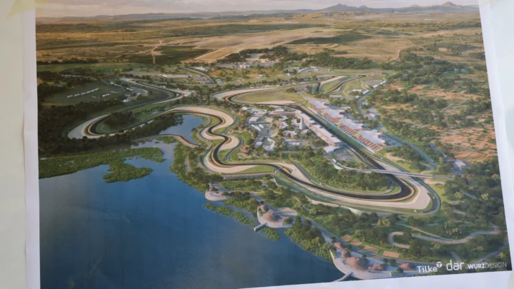 Projeção aérea de um circuito de corrida com curvas sinuosas à beira de um lago, com arquibancadas, prédios de apoio e áreas verdes ao redor