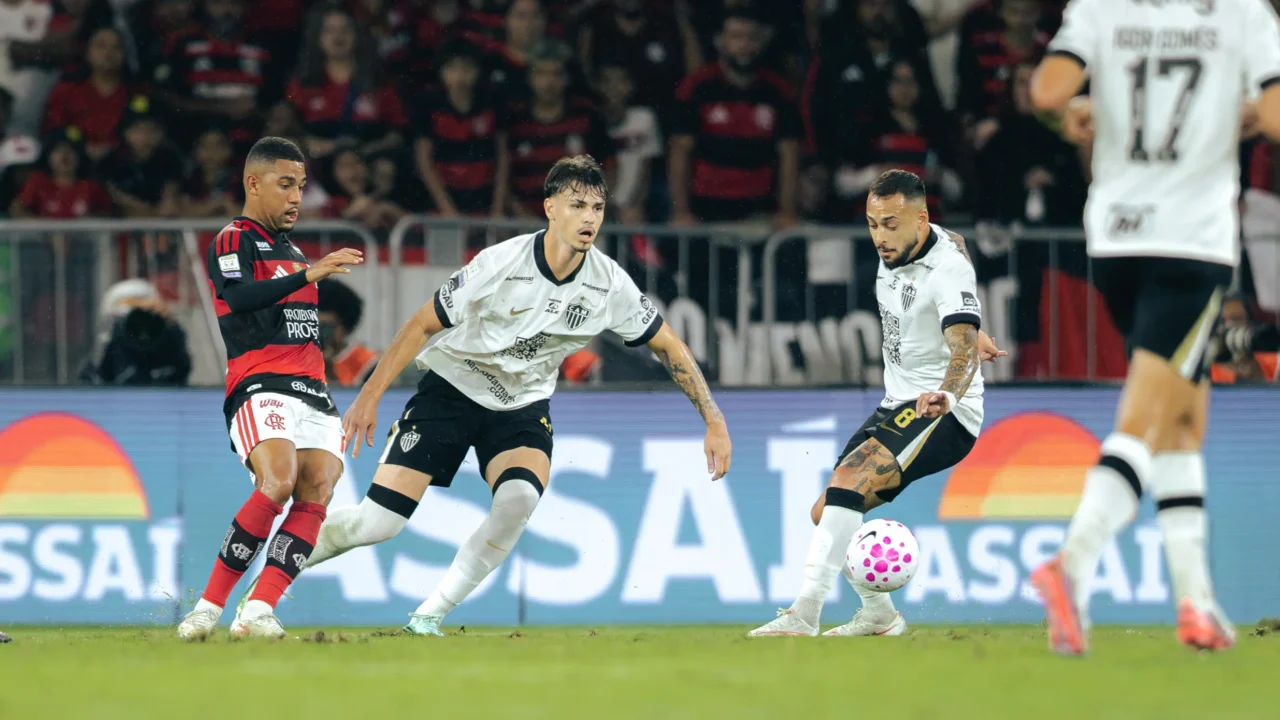 Jogador do Flamengo, de camisa rubro-negra, disputa a bola com dois jogadores do Atlético-MG, de camisa branca, em partida noturna diante da torcida
