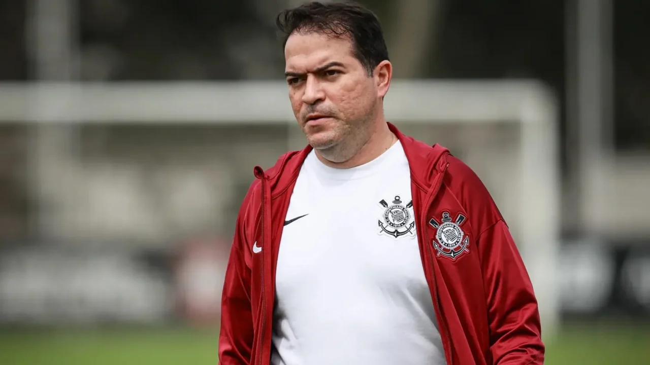 Homem de camisa branca com escudo do Corinthians e jaqueta vermelha, em campo de treino, olhando para o lado