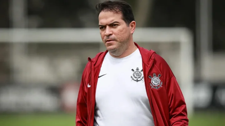 Homem de camisa branca com escudo do Corinthians e jaqueta vermelha, em campo de treino, olhando para o lado