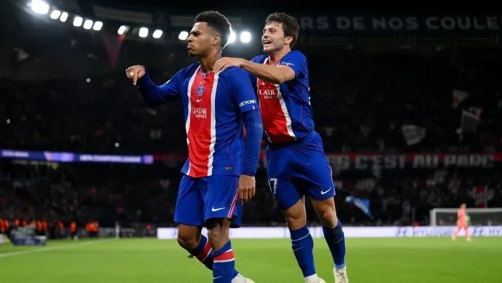 Jogadores do PSG com camisas azul e vermelha comemoram em campo, com um deles apontando para o próprio uniforme, durante jogo da Liga dos Campeões