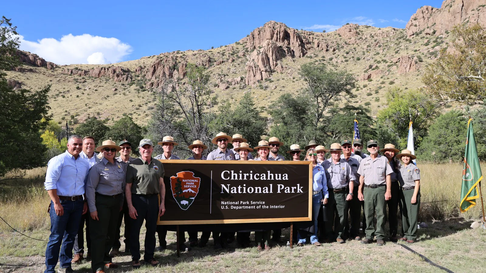 O secretário do Interior, Doug Burgum, o deputado Juan Ciscomani e funcionários do Serviço Nacional de Parques, de chapéu de guarda-parque, posam atrás da nova placa "Chiricahua National Park", com morros de pedra ao fundo