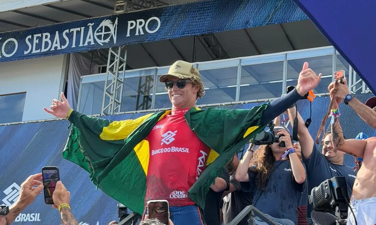 Surfista com bandeira brasileira comemora vitória em pódio de competição, cercado por fotógrafos e torcedores em estádio à beira-mar