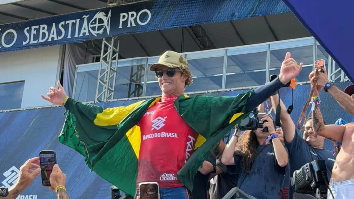 Surfista com bandeira brasileira comemora vitória em pódio de competição, cercado por fotógrafos e torcedores em estádio à beira-mar