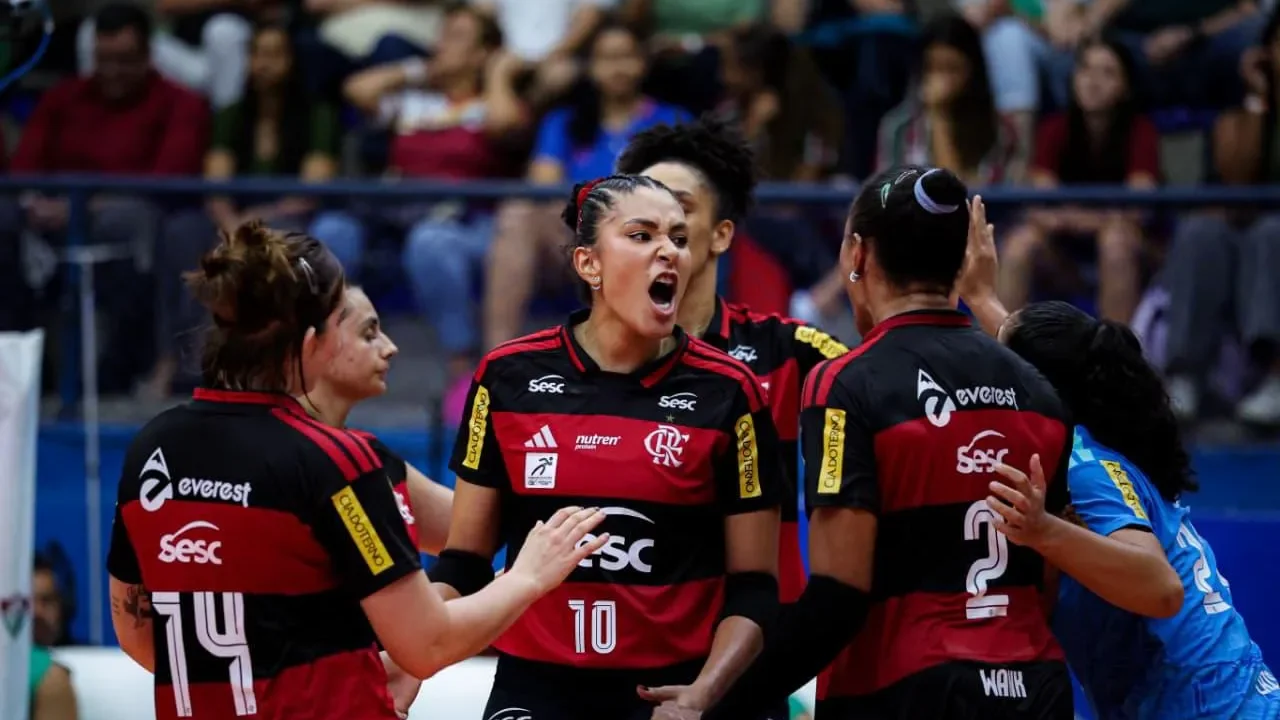 Jogadoras do Sesc-Flamengo comemoram um ponto durante partida de vôlei, com uma atleta de uniforme azul ao lado e torcida ao fundo
