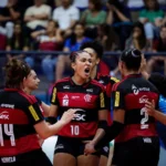 Jogadoras do Sesc-Flamengo comemoram um ponto durante partida de vôlei, com uma atleta de uniforme azul ao lado e torcida ao fundo