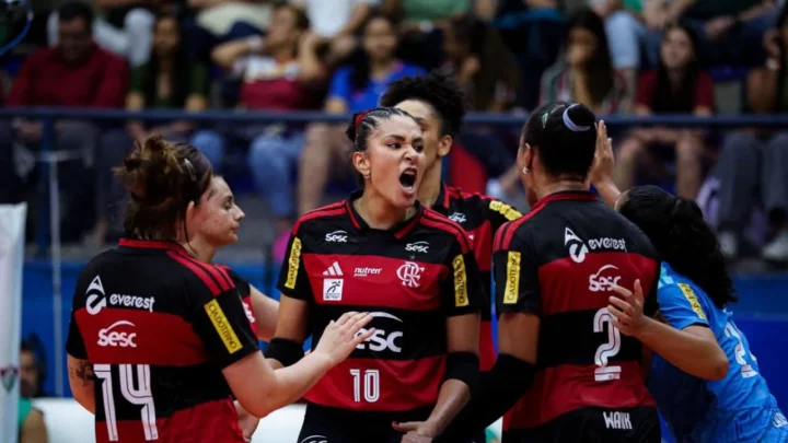 Jogadoras do Sesc-Flamengo comemoram um ponto durante partida de vôlei, com uma atleta de uniforme azul ao lado e torcida ao fundo