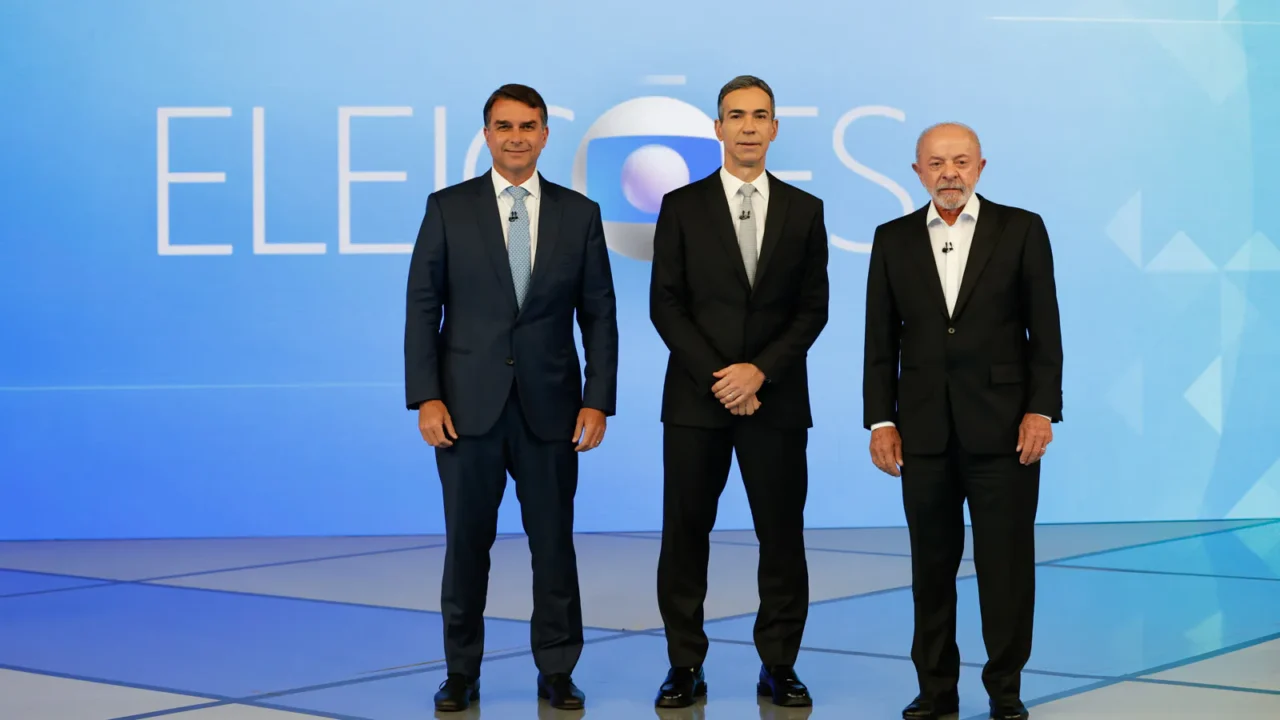Flávio Bolsonaro, à esquerda, e Lula, à direita, de pé no cenário do debate da TV Globo, com o painel "Eleições" ao fundo e um homem de terno preto entre os dois, no Rio de Janeiro, em 23 de outubro de 2026