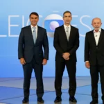 Flávio Bolsonaro, à esquerda, e Lula, à direita, de pé no cenário do debate da TV Globo, com o painel "Eleições" ao fundo e um homem de terno preto entre os dois, no Rio de Janeiro, em 23 de outubro de 2026