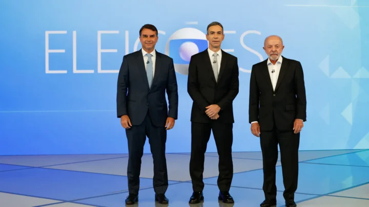 Flávio Bolsonaro, à esquerda, e Lula, à direita, de pé no cenário do debate da TV Globo, com o painel "Eleições" ao fundo e um homem de terno preto entre os dois, no Rio de Janeiro, em 23 de outubro de 2026