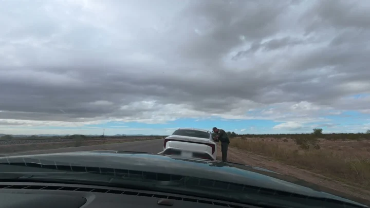 Policial de pé junto à janela do motorista de um carro branco parado no acostamento de uma rodovia, visto a partir do carro estacionado atrás, sob céu nublado