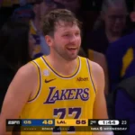 Jogador de camisa amarela e roxa do Lakers, número 77, sorri durante partida, com placar exibido na parte inferior da tela