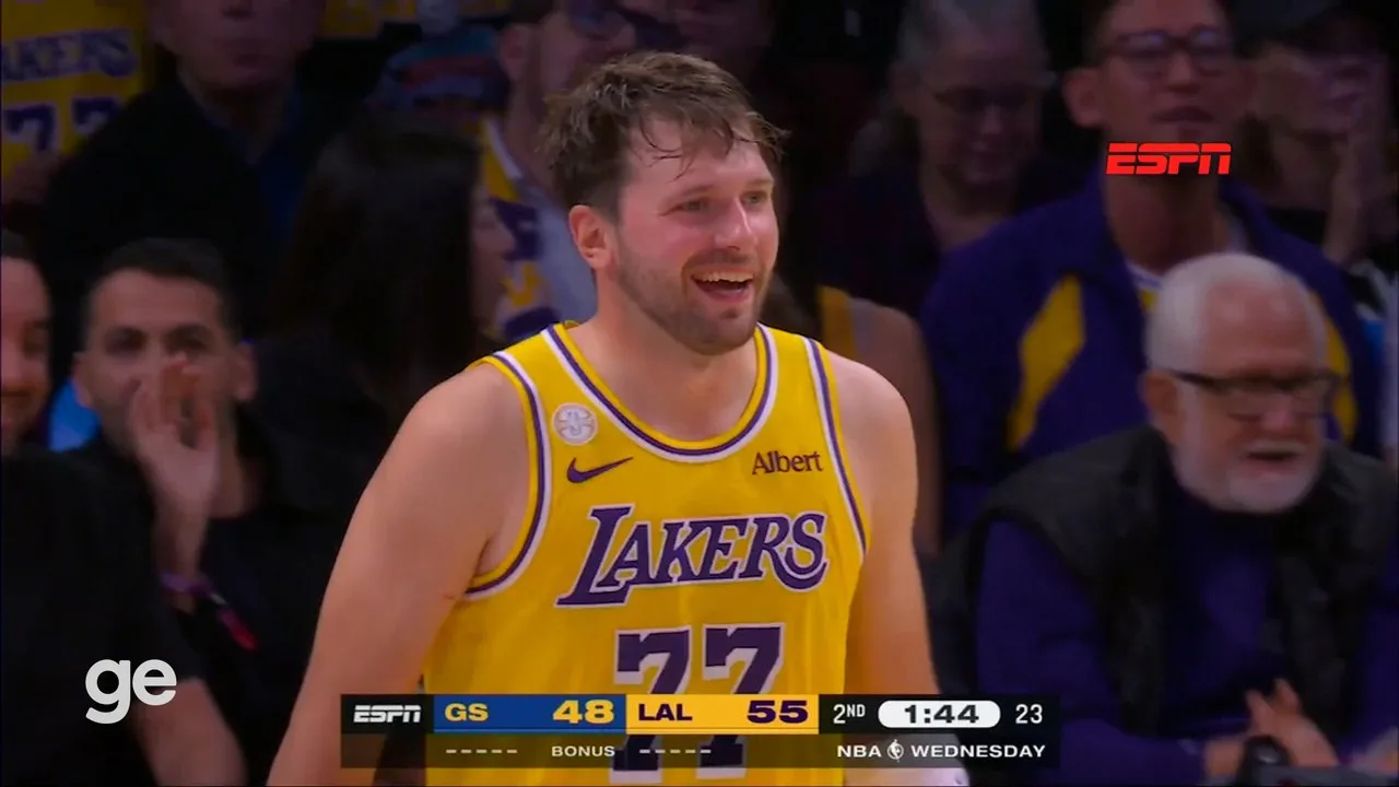 Jogador de camisa amarela e roxa do Lakers, número 77, sorri durante partida, com placar exibido na parte inferior da tela