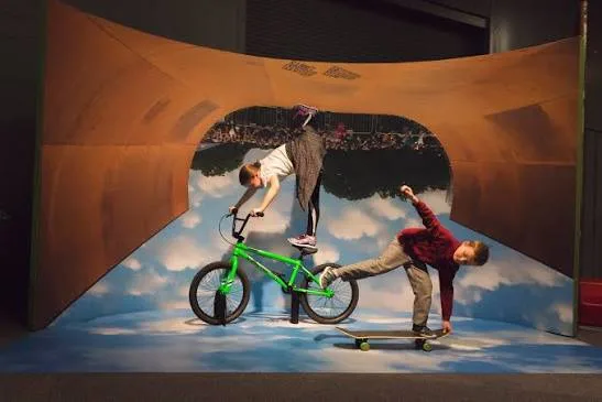 Visitantes interagem com instalação imersiva sobre esportes radicais, com simulação de acrobacias aéreas e bicicleta em ambiente temático do centro de ciências